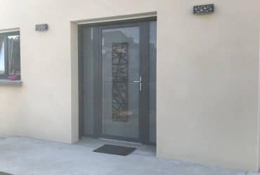 Porte d'entrée vitrée modèle Logan disposant d'un vitrage dépoli, de deux tierces fixes vitrées assorties, le tout dans un coloris Gris anthracite RAL 7016. Elle fut installée avec des fenêtres SO finition extérieure alu 7016 et finition intérieure PVC blanc